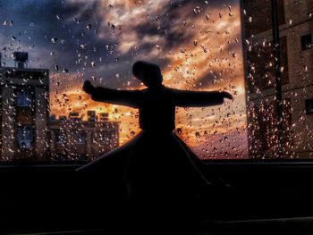 Silhouette man by glass window against building during sunset