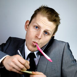 Close-up portrait of businessman talking on phone while brushing teeth against gray background