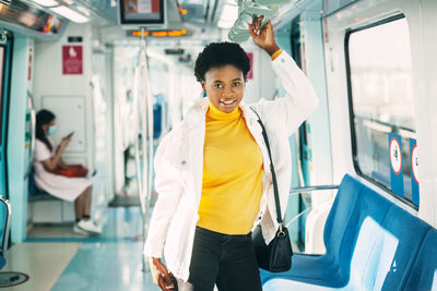 A smiling young african woman holds on to a handrail while standing on a subway 