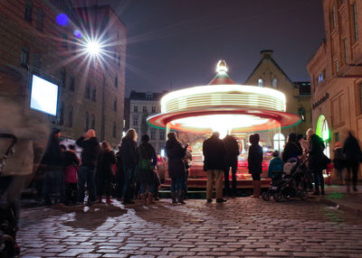 Rear view of people standing in front of spinning carousel at night