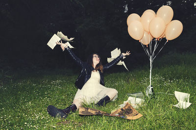 Full length of happy woman throwing papers while sitting by balloons on grassy field during dusk