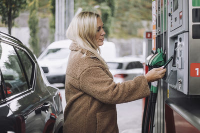 Side view of blond woman doing payment via smart phone while standing at fuel station