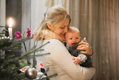 Side view of mother carrying baby girl by christmas tree at home