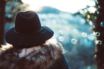 Rear view of woman wearing hat and fur coat while looking at bubbles in mid-air