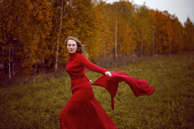 Woman in a red dress and sweater stands in a field with trees in autumn