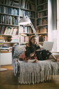 Mother using laptop while down syndrome daughter with digital tablet on sofa in living room