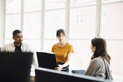 Female software engineer explaining to coworkers while sitting against window in office