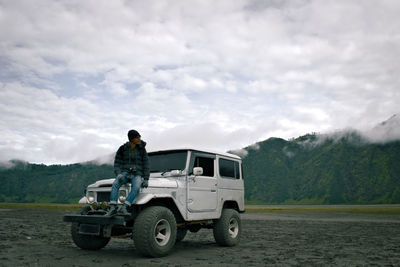 Full length of hiker sitting on off-road car against cloudy sky at landscape