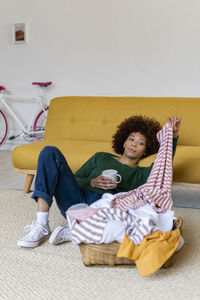 Bored woman holding t-shirt and coffee mug reclining on sofa at home