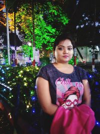 Portrait of young woman standing against trees at night