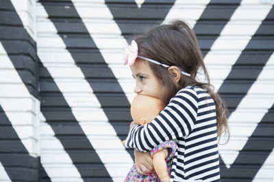 Cute girl looking away while holding doll against wall