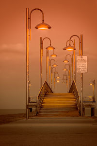 Illuminated street light over walkway at sea shore against sky during sunset