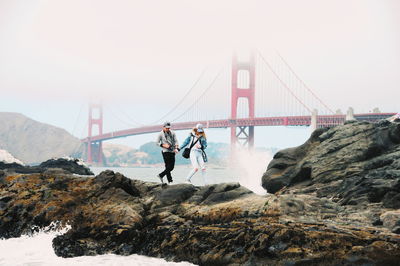 Full length of couple standing against suspension bridge