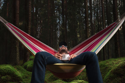 Man having drink while relaxing on hammock in forest