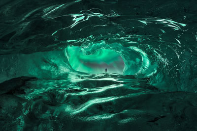Through hole in ice cave view of explorer standing under night sky with bright green aurora borealis in lofoten islands