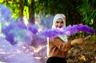 Portrait of cheerful young woman holding purple distress flare while standing against trees