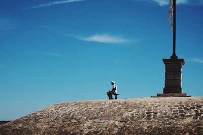 Low angle view of mature man sitting on bench against blue sky during sunny day