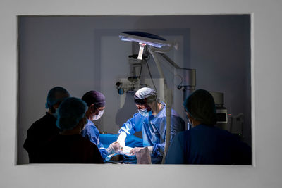 Through glass unrecognizable surgeons and nurses in uniform concentrating and operating patient using special equipment in operating room of contemporary hospital
