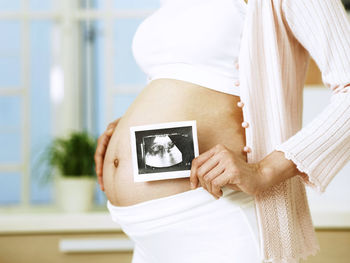 Midsection of pregnant woman holding photograph while standing indoors