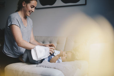 Smiling mother dressing baby boy on sofa at home