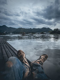 Low section of man relaxing on pier over lake against cloudy sky