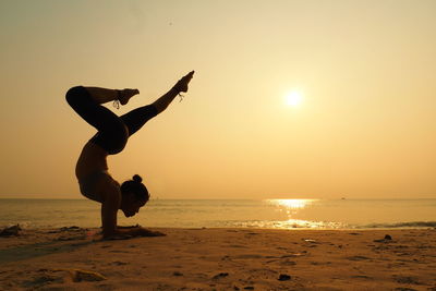 Full length of woman practicing yoga on shore at beach against sky during sunset