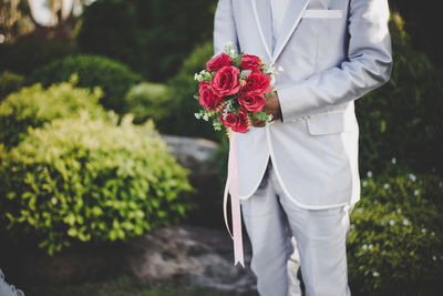 Midsection of bridegroom holding rose bouquet while standing outdoors