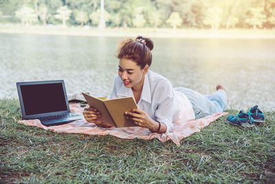 Smiling woman reading book while lying by laptop at lakeshore 