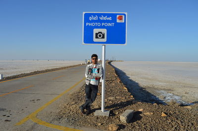 Portrait of young man standing on road by sign board against sky