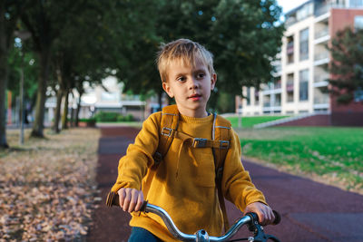 A boy in a yellow sweater with a yellow backpack rides a bicycle in the park.