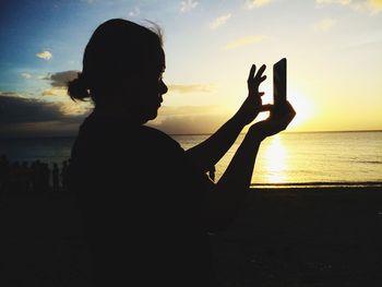 Silhouette woman photographing on mobile phone at beach against sky during sunset