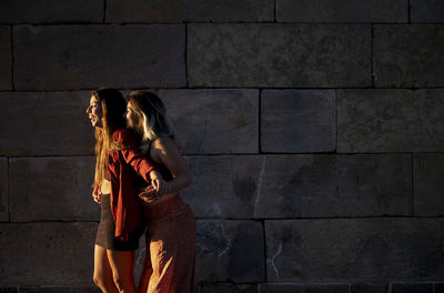 Two girls standing against stone wall during sunset