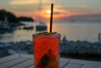 Red cocktail, focus on foreground, sunset at sea in background.