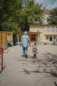 Grandfather with granddaughter walking on footpath towards kindergarten