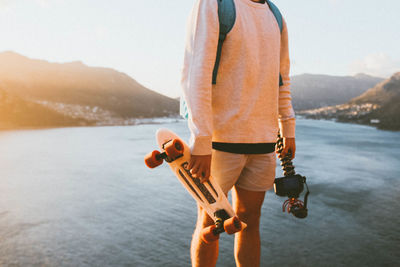 Midsection of man holding skateboard and camera against lake