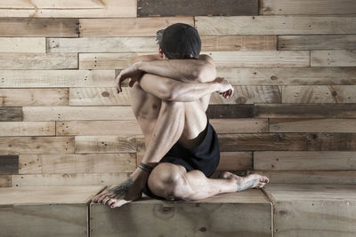 Rear view of shirtless man sitting on wood