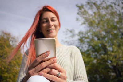 Low angle view of young woman using mobile phone while sitting against sky at park