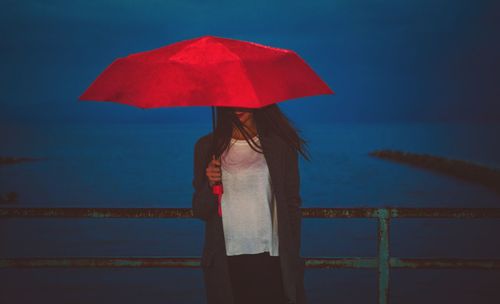 Woman standing with red umbrella by railing on pier against sea