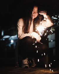 Young woman holding illuminated sparkler while crouching at night