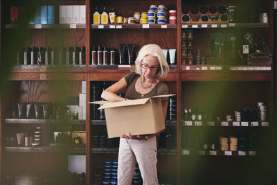 Senior saleswoman unpacking cardboard box against rack in deli