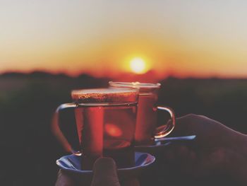 Cropped hands holding black coffee cup against sky during sunset