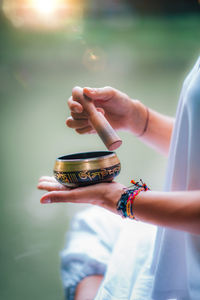 Midsection of woman holding singing bowl while doing yoga at lake