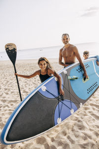 Shirtless man carrying surfboard with daughter and son while walking on sand at beach