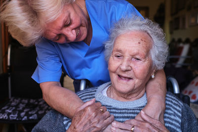 Caring nurse in blue uniform embracing elderly disabled woman with gray hair sitting in wheelchair in light room at home