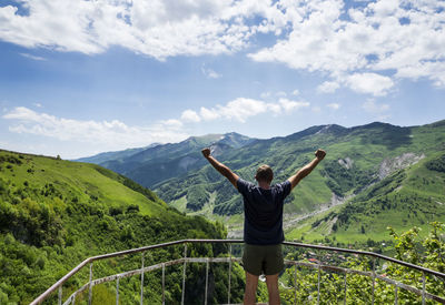 Man with arms raised standing at observation point against mountain and cloudy sky