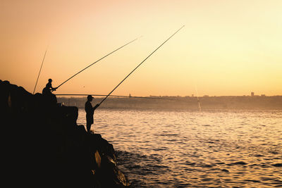 Silhouette fishing rod by sea against sky during sunset