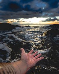 Close-up of hand over sea against sunset sky