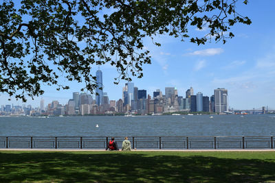 Rear view of people sitting at park by hudson river and city skyline against sky