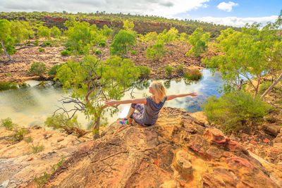 Rear view of woman with arms outstretched sitting by river on rock at kalbarri national park 
