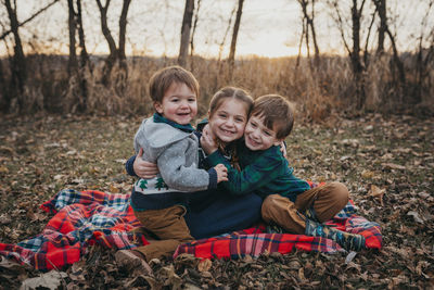 Three siblings sitting on blanket hugging smiling at camera